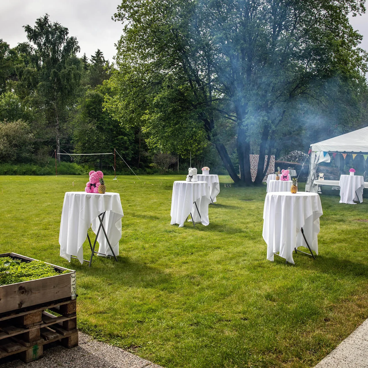 Round tables with white tablecloths display pink and white flower arrangements. A large tent and trees provide a scenic backdrop in a grassy outdoor event setting.