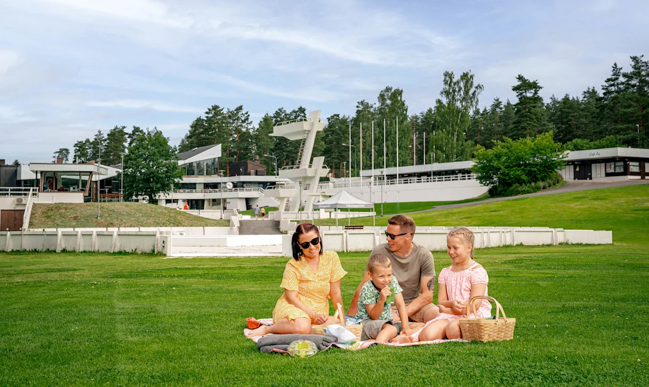 Perhe istuu piknikhuovalla, syö ja keskustelee. Heitä ympäröi laaja vihreä nurmikko, jossa on puita ja moderneja rakennuksia taustalla.