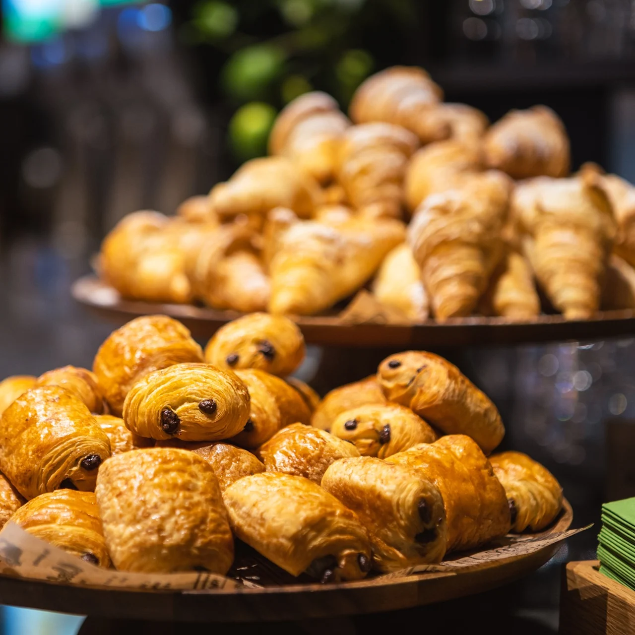 Bakverk visas på träfat i ett bageri; chokladcroissanter är staplade i förgrunden, med vanliga croissanter i bakgrunden. Gröna servetter är prydligt staplade i närheten.