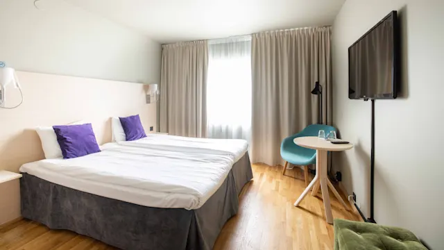 En prydligt bäddad enkelsäng med vita lakan och lila kuddar är placerad nära ett stort fönster med gardiner. Ett litet träbord och en blå stol är placerade bredvid under en väggmonterad TV.
