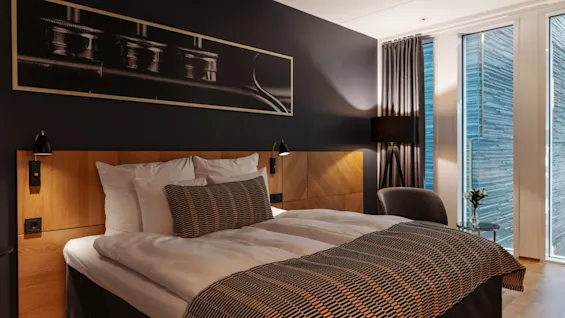 Et moderne og komfortabelt hotellrom med en stor seng, sengegavl i tre, mørk aksentvegg og lyse vinduer.