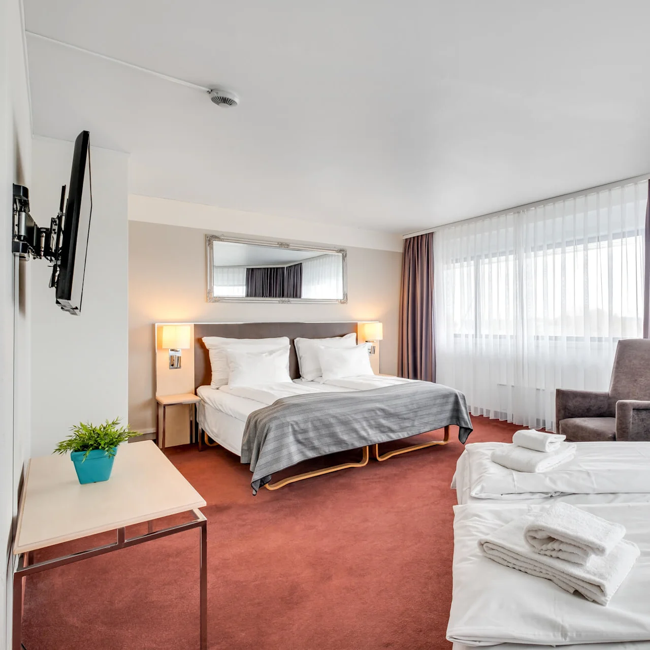 A hotel room with two neatly made beds, modern furniture, and large windows draped with sheer and opaque curtains.