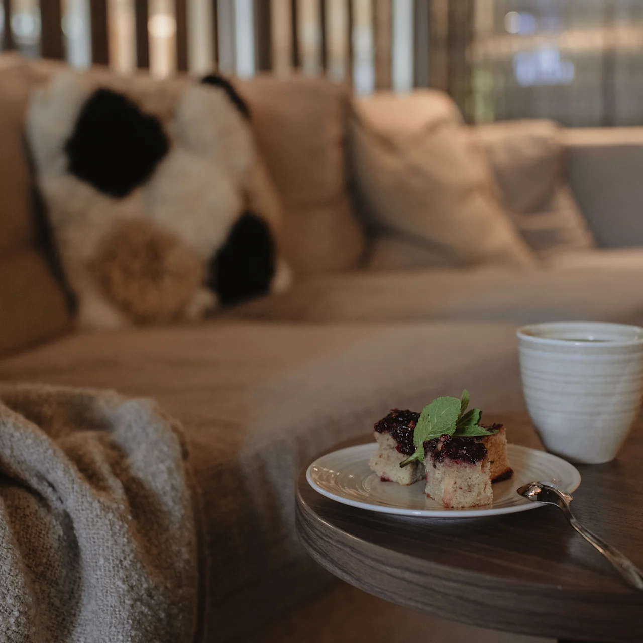 A plate with berry-topped cake sits on a small table beside a cup. The setting includes a cozy sofa with pillows and a blanket.