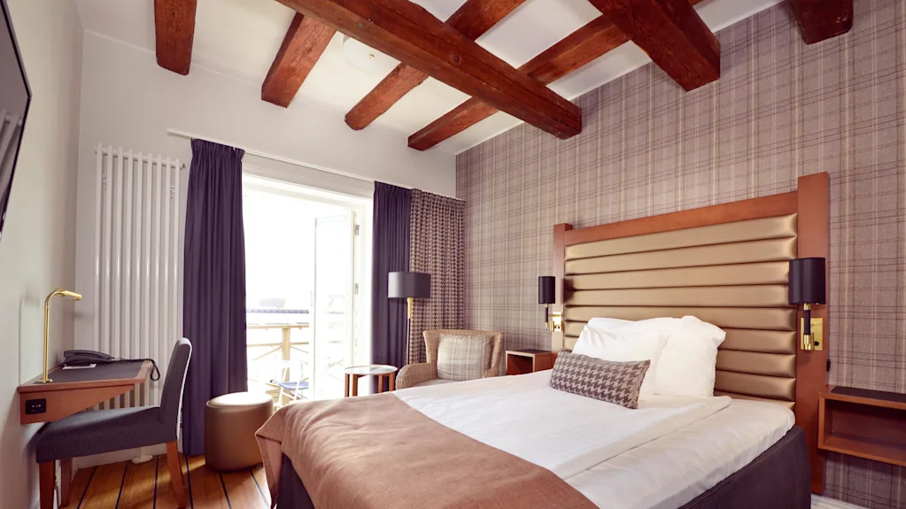 A tidy bedroom with a made bed, wooden desk, and chair, set in a room with wooden ceiling beams and a balcony.