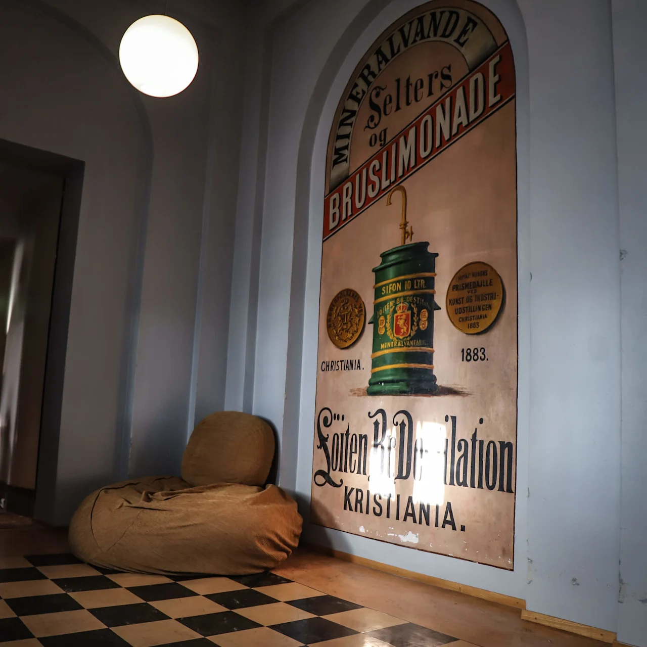 A large vintage advertisement decorates a wall above a cushioned chair in a room with a checkered floor. Text reads: "Mineralvande Selters og Bruslimonade... Løiten Brænderi-Kr Prøve."