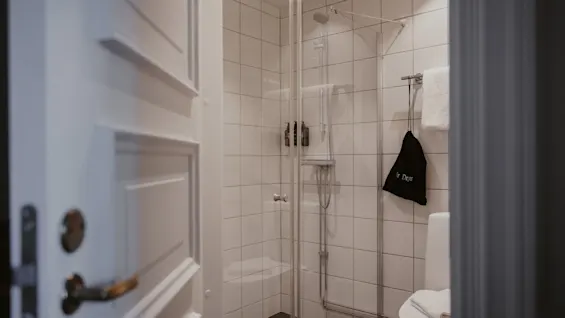 Ett rent hotellbadrum med en glasdusch, vitkaklade väggar och en toalett. En svart väska märkt Hair Dryer hänger bredvid en handduk.