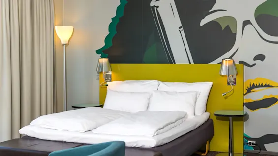 A bed with white linens is set against a yellow headboard. Two wall lamps provide light. A mural features a stylized face with sunglasses in the background.