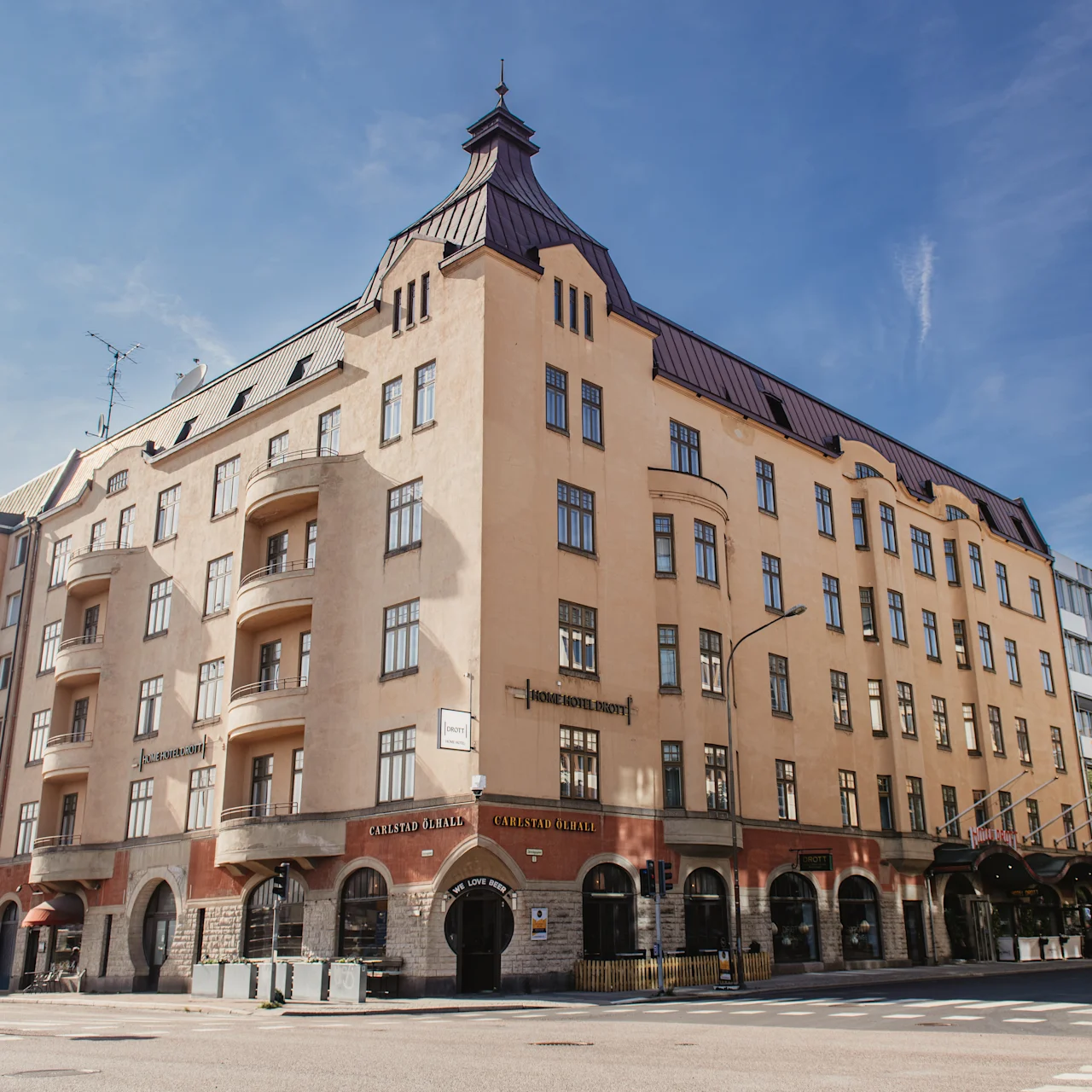 En beige fler-etagers bygning står på et gadehjørne med bueformede vinduer, altaner og et stejlt tag. Et træ og andre bygninger omgiver det. Teksten lyder "Fennia Salonki" og "Fennia Restaurant".