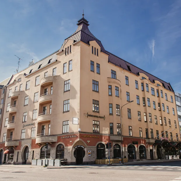 Ett beige fler-våningshus står på ett gatuhörn med välvda fönster, balkonger och brant tak. Ett träd och andra byggnader omger det. Texten lyder "Fennia Salonki" och "Fennia Restaurant".