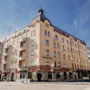 Ett beige fler-våningshus står på ett gatuhörn med välvda fönster, balkonger och brant tak. Ett träd och andra byggnader omger det. Texten lyder "Fennia Salonki" och "Fennia Restaurant".