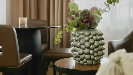 A textured vase with green and purple foliage sits on a round table in a cozy living room with brown chairs and curtains.