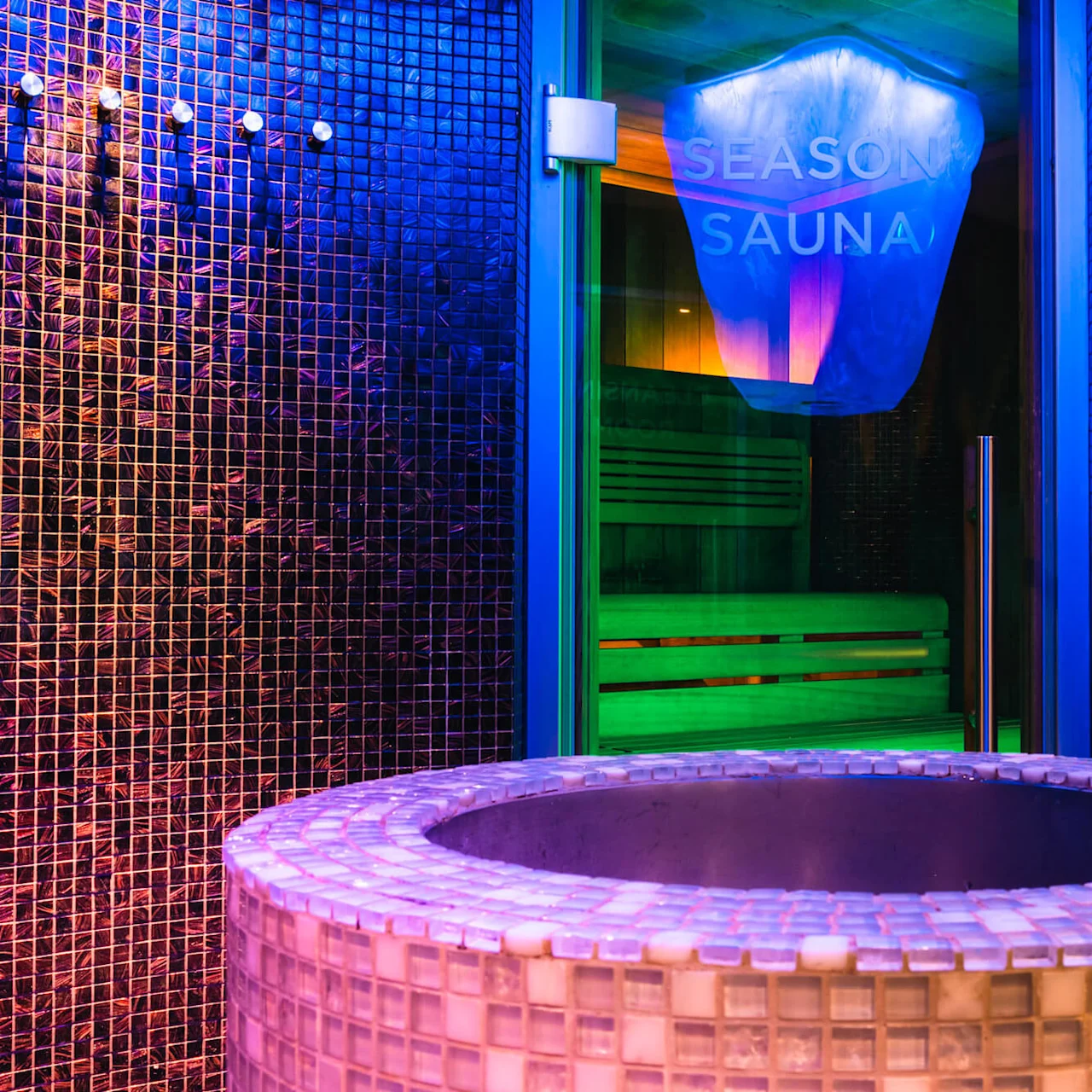 Ett mosaikbelagt runt basin står framför en glasdörr märkt "Season Sauna." Neonlys skapar en livlig atmosfär med synliga träbänkar inuti.