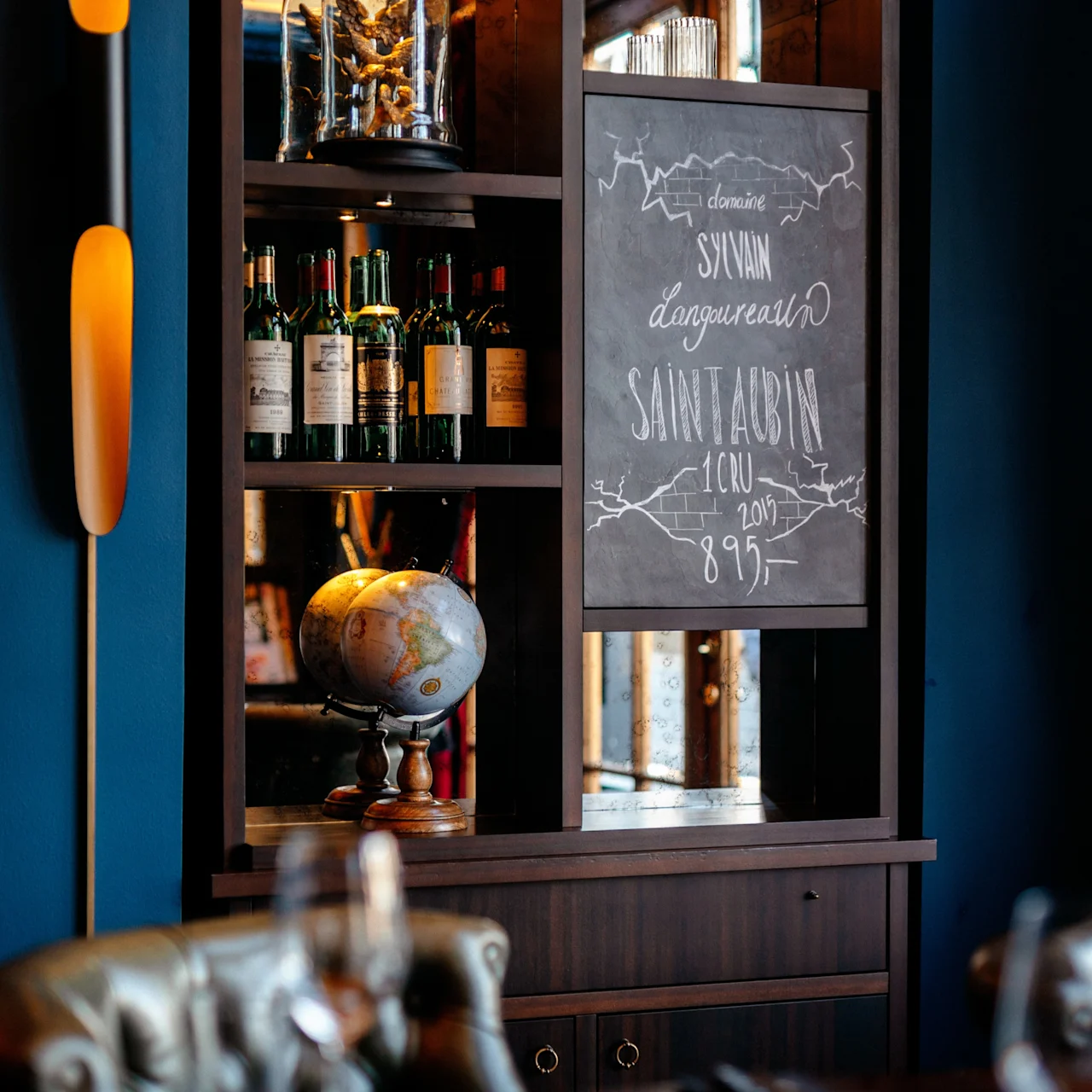Shelf holds wine bottles and a chalkboard with text: "domaine Sylvain Langoureau Saint Aubin 1er cru 2017 895". A globe sits below. Dining table in foreground.