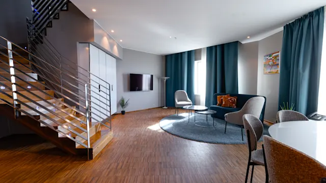 A modern living room with a stairway on the left and a seating area on the right, featuring a sofa, chairs, and a wall-mounted TV. Natural light enters through large windows.