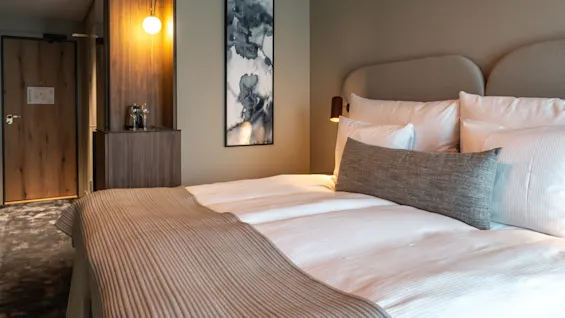 En prydligt bäddad säng med vita lakan och en beige kastfilt står i ett hotellrum. Ett träskåp och abstrakt konst pryder väggen i bakgrunden, med mjuk belysning.