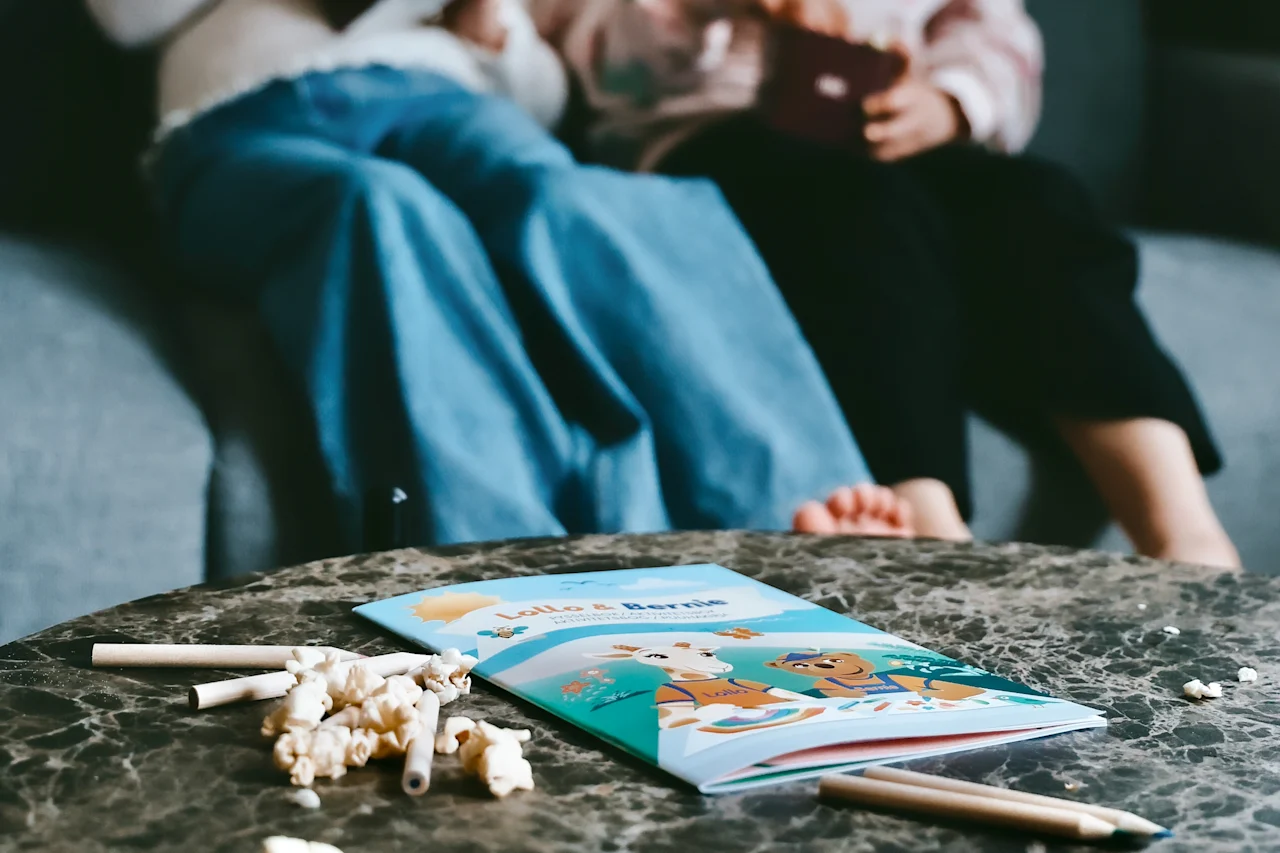 Coloring book with cartoon animals lies open on a marble table, surrounded by scattered popcorn and pencils. Two people sit on a sofa in the background.