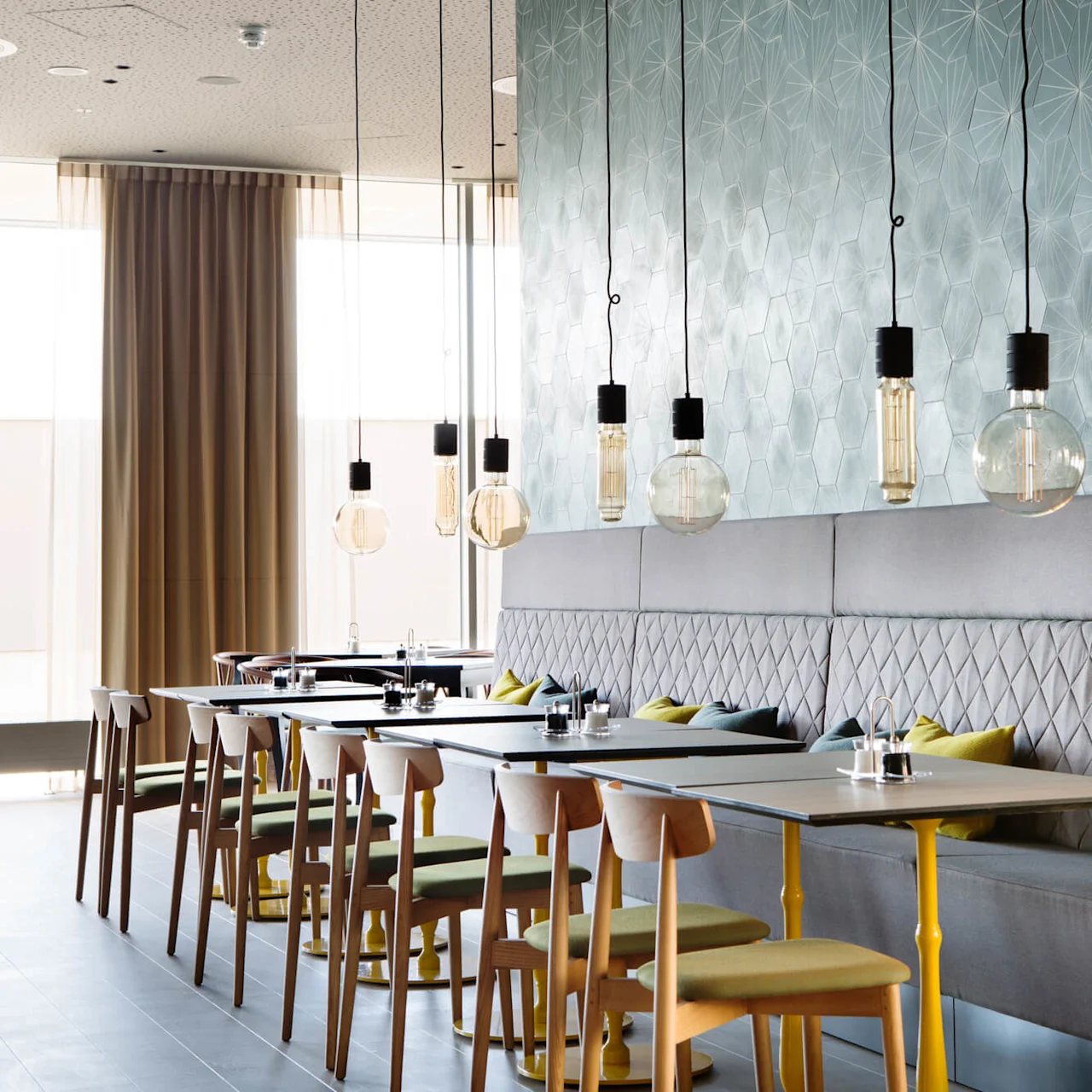 Pendant lights hang above a row of tables with wooden chairs and cushioned benches. Geometric patterned wall and large windows create a modern restaurant atmosphere.