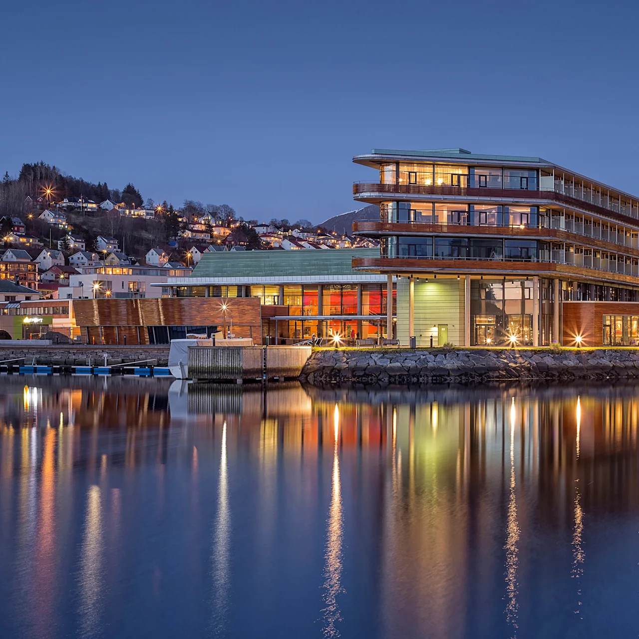 En opplyst moderne bygning står ved rolig vann, reflekterer sine lys. Trappede terrasser og store vinduer kjennetegner den, med en by og åser i bakgrunnen under en klar kveldshimmel.