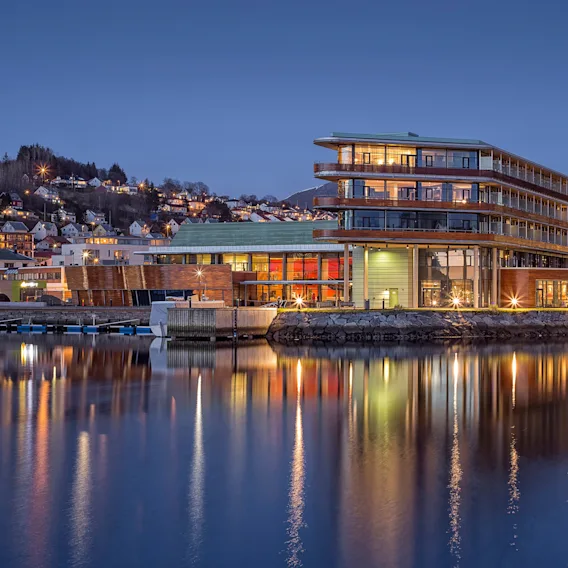 En opplyst moderne bygning står ved rolig vann, reflekterer sine lys. Trappede terrasser og store vinduer kjennetegner den, med en by og åser i bakgrunnen under en klar kveldshimmel.