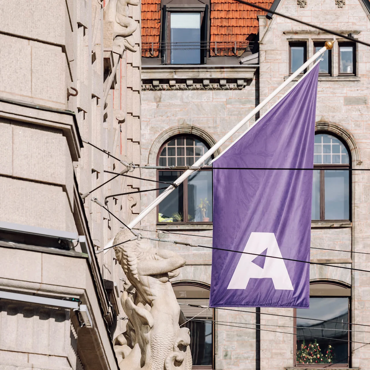 Purple flag with a large white letter "A" hangs from a building. Background features a stone facade with several arched windows and red roof tiles.