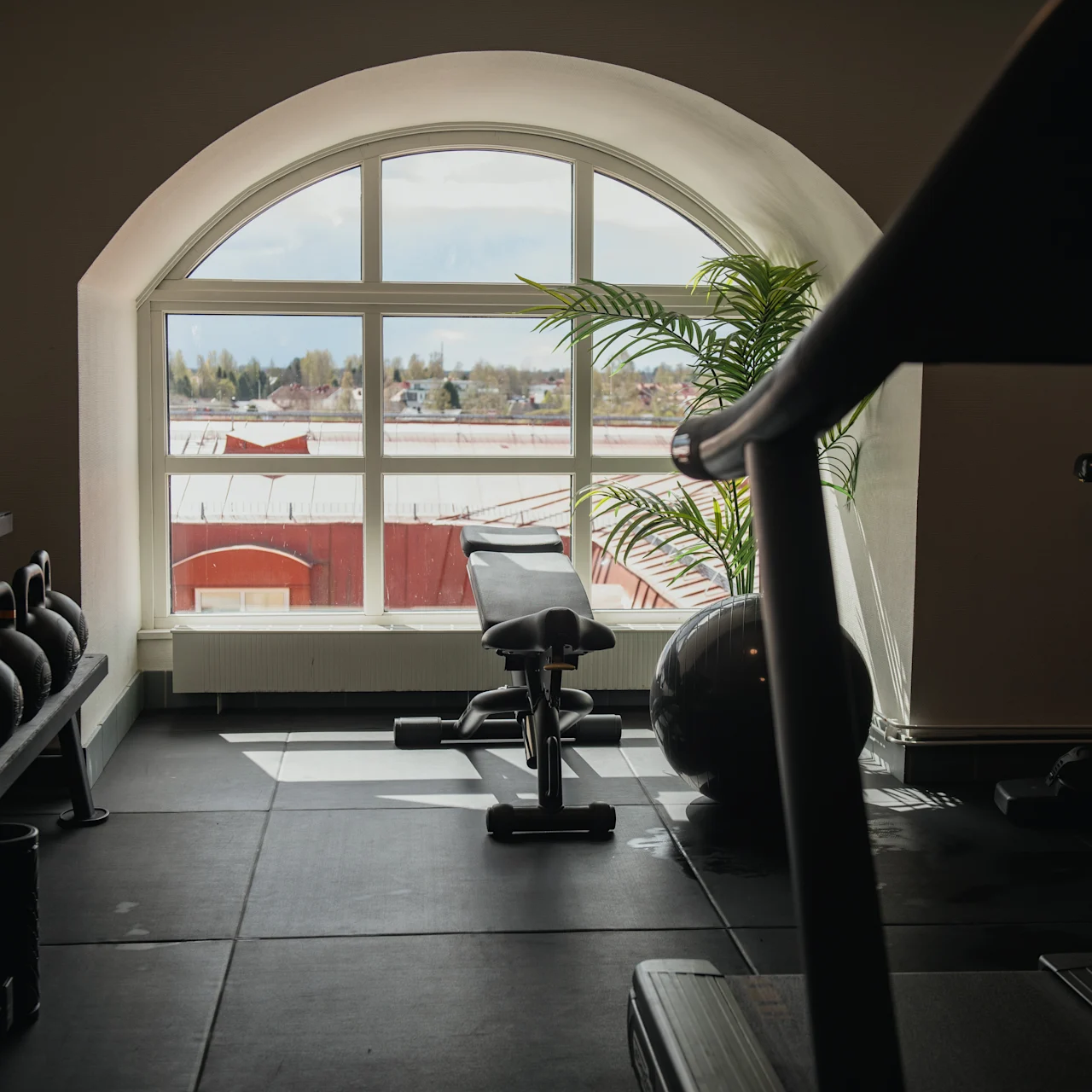A gym bench faces a large arched window in a fitness room. Dumbbells and exercise equipment surround it, with a view of rooftops outside.