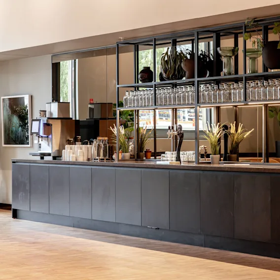 A sleek black counter holds coffee machines, cups, and decorative plants, set in a modern, open space with a mirrored backdrop displaying bottles and greenery.
