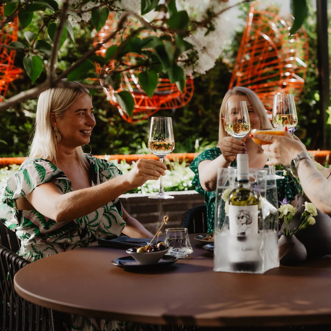 Four people toasting with wine glasses at an outdoor table, surrounded by green plants and orange decorative elements. The table holds a wine bottle in an ice bucket and small dishes.