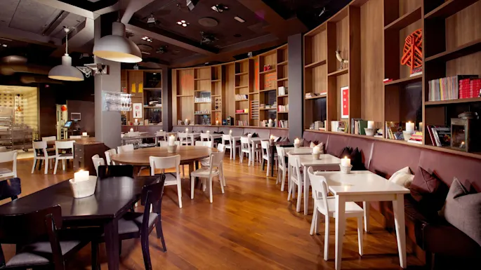 A modern, warmly-lit café interior with wooden floors, white and dark chairs, and tables with candles, against a backdrop of large bookshelves.