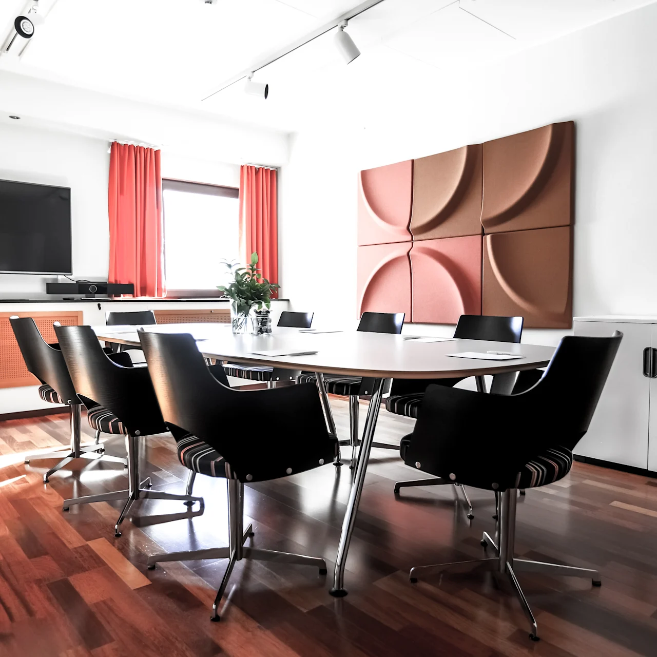 Conference table surrounded by chairs sits in a modern meeting room, with a TV on the wall and red curtains framing a window. Wooden flooring adds warmth to the space.