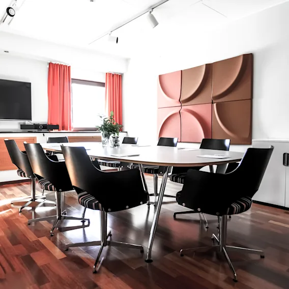 Konferensbord omgivet av stolar står i ett modernt mötesrum, med en TV på väggen och röda gardiner som ramar in ett fönster. Trägolv ger värme till rummet.