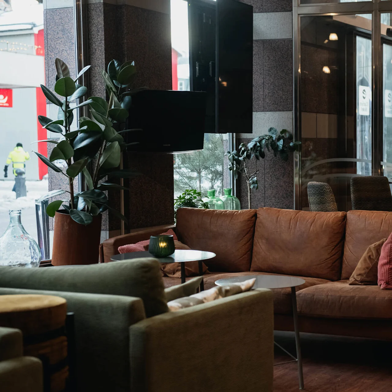 Brown leather sofa in a cozy lounge area, surrounded by green plants and candles on tables, located indoors near large windows with a view of a snowy street.