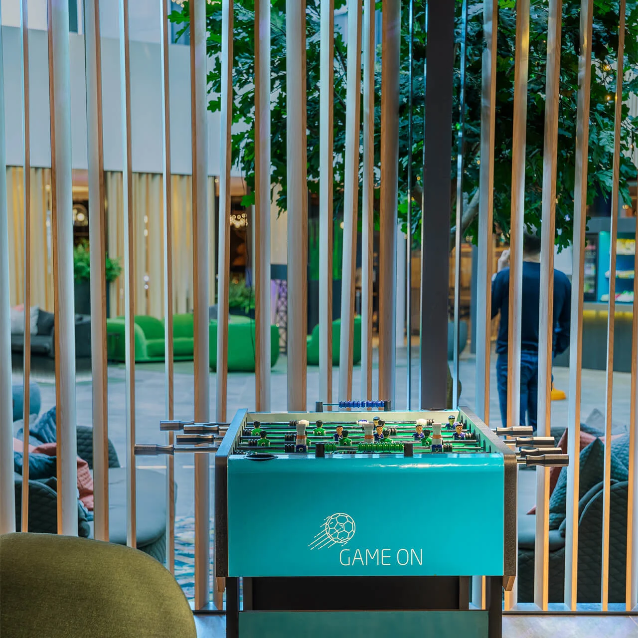 Foosball table stands stationary, green and labeled "GAME ON," situated indoors with a modern seating area and wooden slats in the background.