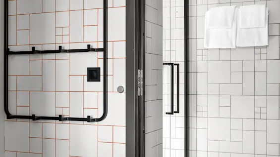 Ett badrum har geometriskt kaklade väggar i vitt med orange och svarta linjer, en svart metallhanddukshållare och två vita handdukar nära en glasdörr med svarta handtag.