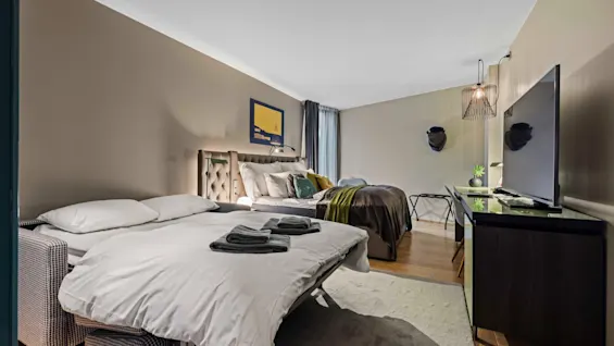 Bed standing with neatly folded towels, near a larger bed, in a cozy, modern bedroom. Features hardwood floors, a TV on a stand, wall art, and light fixtures.