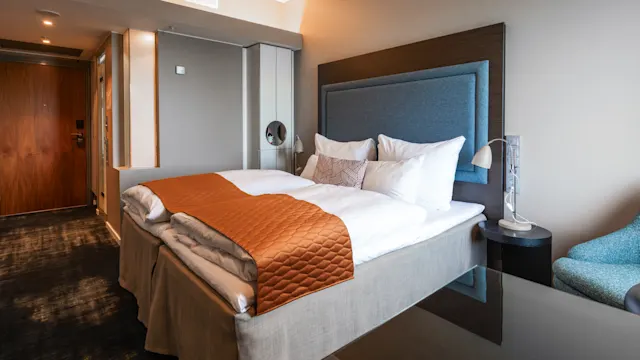 Voitää hotellihuone modernissa tyylissä, jossa on siististi pedattu sänky, valkoiset tyynyt ja oranssi peitto. Vieressä yöpöytä, valaisin sekä puuovi ja matto.