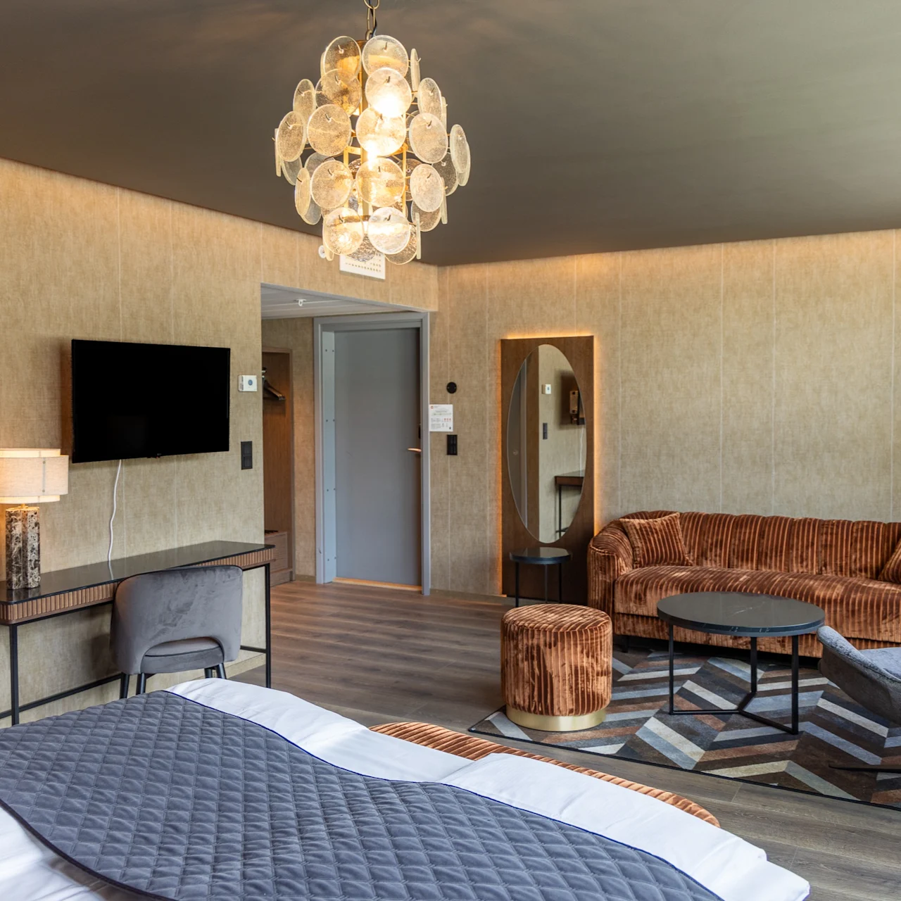 Ett hotellrum har en säng med ett grått quiltat täcke, en mysig sittgrupp med orange möbler och en platt-TV på väggen. Modern inredning kompletterar den varma belysningen.