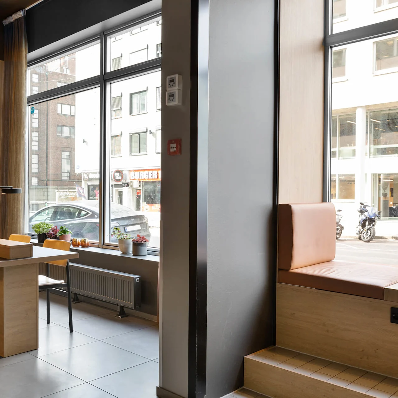 A modern workspace with a long table and chairs stands beside large windows. Neon text on the wall reads, "WORKING CLASS HERO." Outside are buildings and a parked car.
