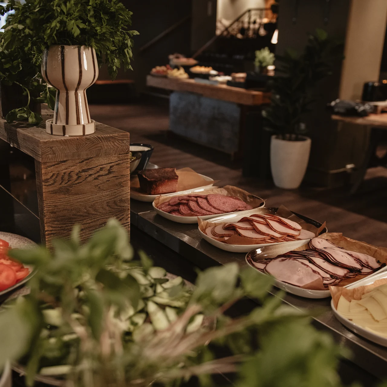 Plates of sliced meats, cheeses, and vegetables are arranged on a wooden buffet table, with greenery in the foreground and other tables visible in the background.