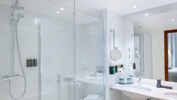Brusekabine med glasdør i moderne hvidflisebelagt badeværelse. I nærheden er der toilet og håndvask med stort spejl. Blødt lys fremhæver den rene, minimalistiske stil.