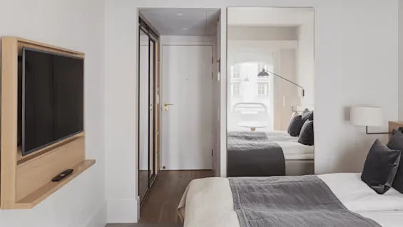 Television mounted on a wooden frame, displaying a blank screen, adjacent to a modern hotel room with a bed and large mirror reflecting a window. A remote rests below.