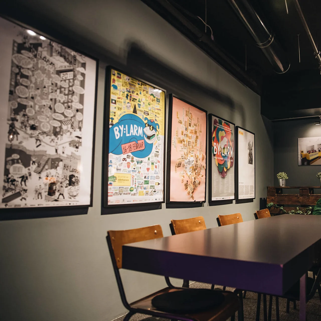 Tables and chairs sit in a dimly lit room, surrounded by six framed artworks on a green wall, including text reading "BY:LARM 16-18 FEBRUAR." Two small plants rest on a wooden shelf.