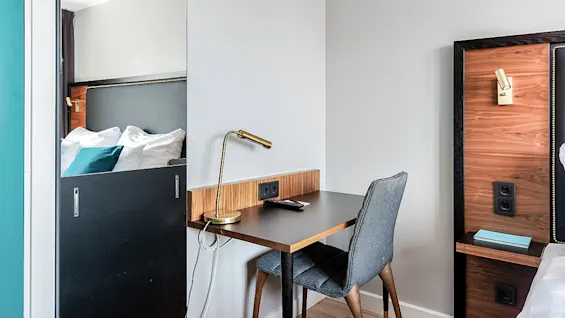 A modern hotel room with an open door revealing a bed, a desk with a lamp, chair, and wooden wall panels.