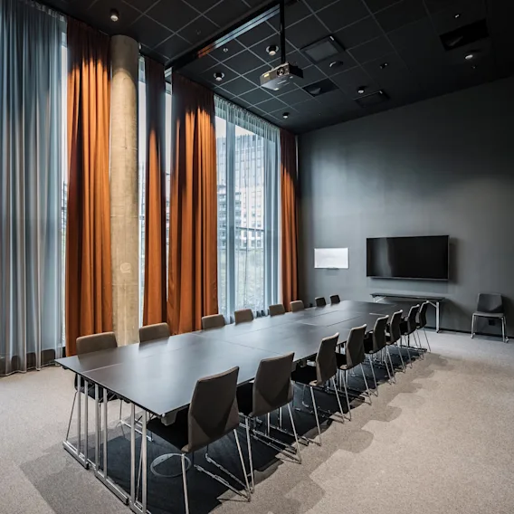 Et langt konferansebord med stoler står i et moderne møterom, med høyt tak, store vinduer med oransje gardiner og en veggmontert TV.
