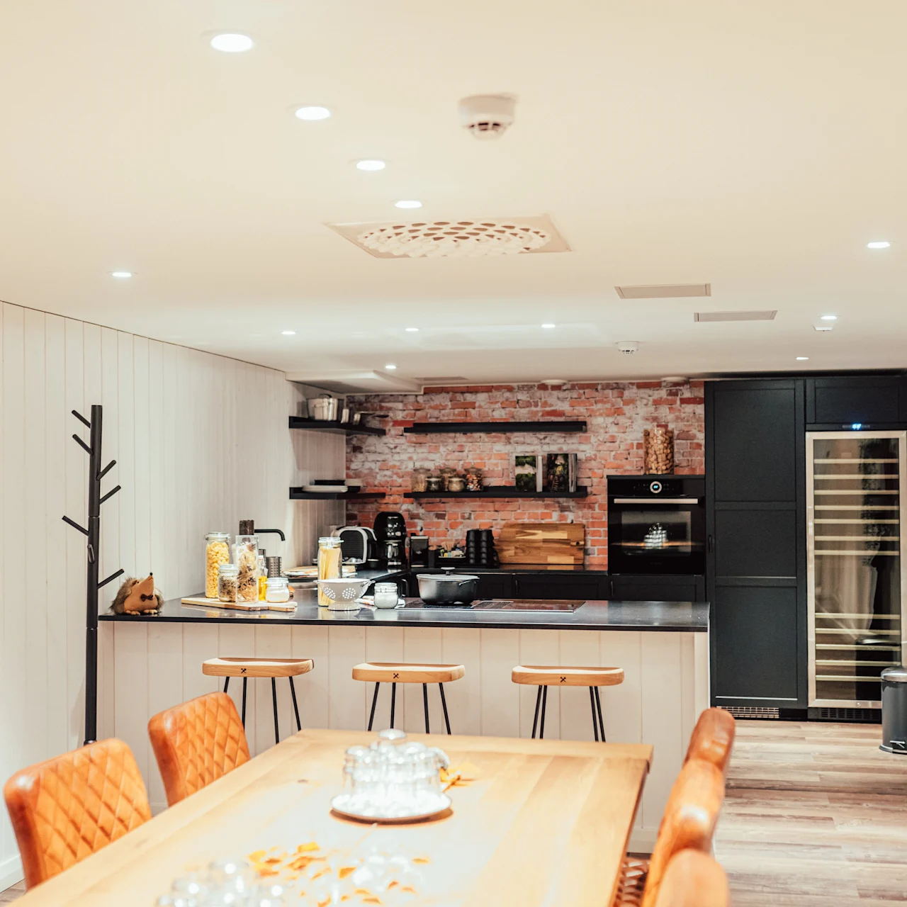 A modern kitchen with a dining table and orange chairs, featuring a counter with stools, appliances, and decorated with exposed brick and wood. Bright overhead lights illuminate the space.