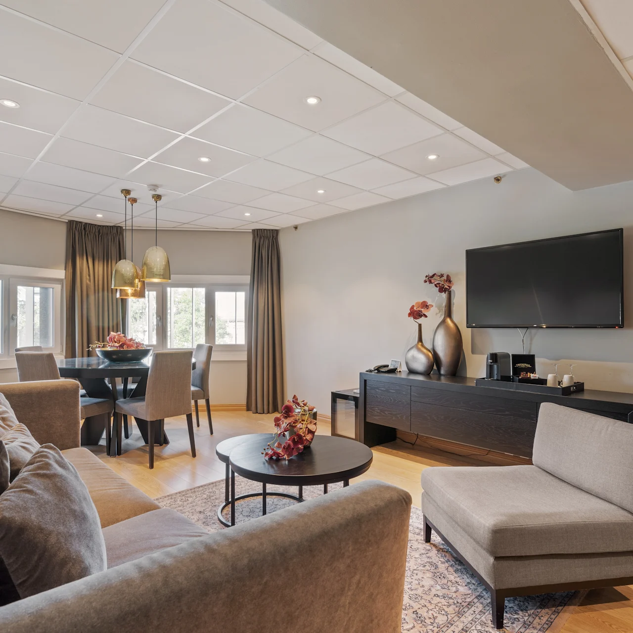 A living room with a gray sofa and chair sits next to a coffee table with flowers; a dining area with windows and pendant lights is nearby, while a TV hangs on the wall.