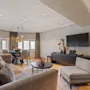 En stue med en grå sofa og stol står ved siden af et kaffebord med blomster; et spiseområde med vinduer og pendellamper er i nærheden, mens et TV hænger på væggen.
