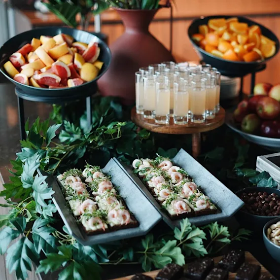 Et buffetbord viser et udvalg af fødevarer, herunder frugtfade, rejeåbne sandwiches, nødder, små desserter i glas og æblebunker, dekorativt arrangeret med grønne blade.