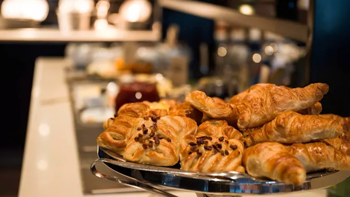 Croissants and pastries rest on a silver platter; the background is a blurred kitchen counter with various jars and containers.