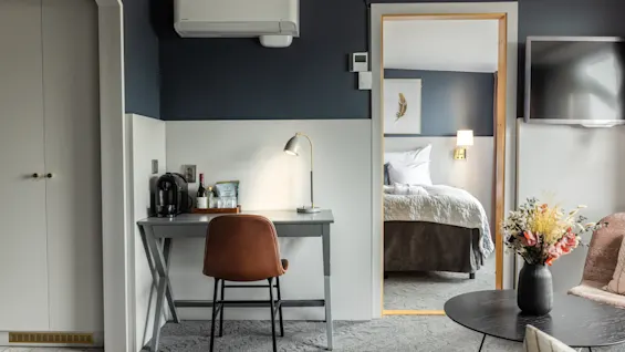 Ett mysigt rum med ett enkelt skrivbord och kaffemaskin, som leder till en snyggt bäddad säng i ett angränsande rum. En bukett blommor ger en varm känsla till den moderna inredningen.