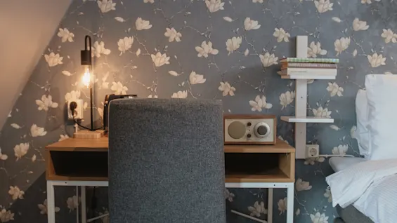 A desk with a lamp and a radio sits against a floral-patterned wall. A gray chair faces the desk in a cozy bedroom setting.